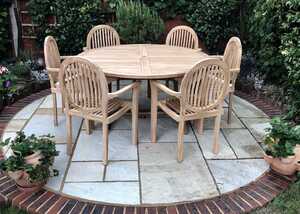 Round teak table with five teak chairs on a stone patio surrounded by greenery.