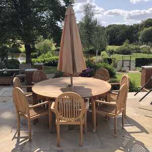 Teak outdoor dining table with a large parasol and surrounding teak chairs, set in a scenic garden.