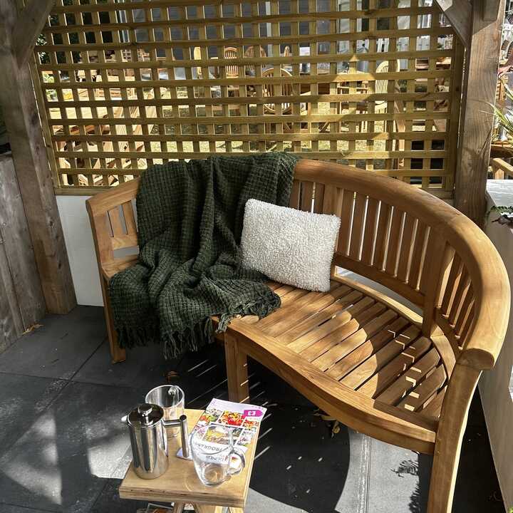 A teak bench with a white cushion, a green blanket and a small table holding a coffee pot and magazines.