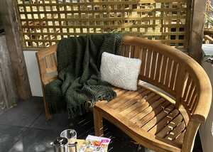 Teak garden bench with a green blanket and white cushion, small table with coffee and magazines.