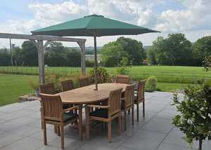 A dining area with a teak wooden table and teak chairs under a green parasol, in a grassy field.